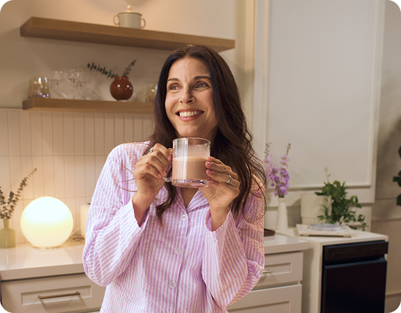 Smiling woman in pink striped pajamas holding a mug of creamy sleep beverage in a warmly lit kitchen